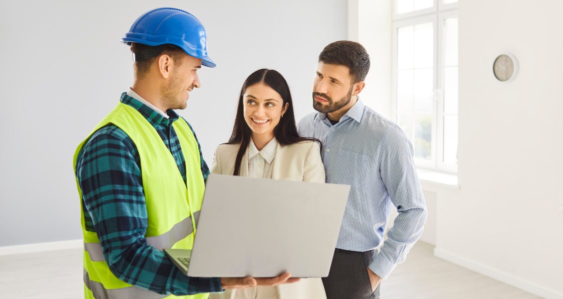 Contractor in a hard hat walks two clients through a bid proposal on a laptop