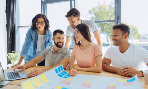 Five colleagues gather around a laptop and a sheet of sticky notes to plan sales battle cards