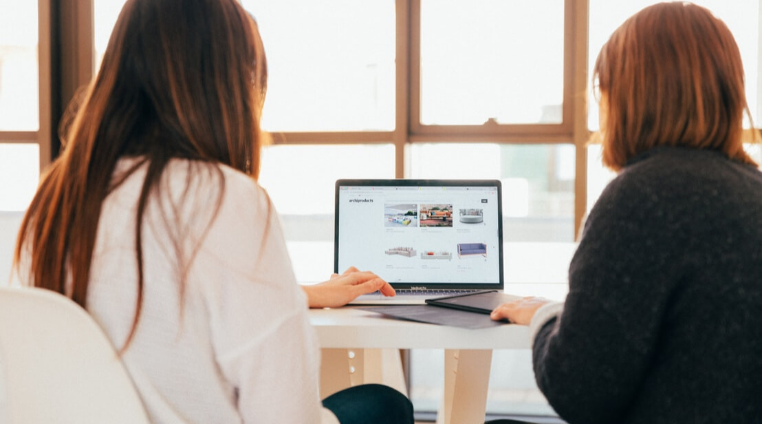 Two women sitting at a desk looking at a laptop screen.