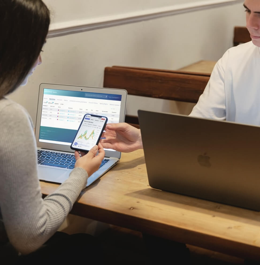 A woman and a man sitting opposite eachother at a desk with a laptop each and both looking at a mobile phone screen.